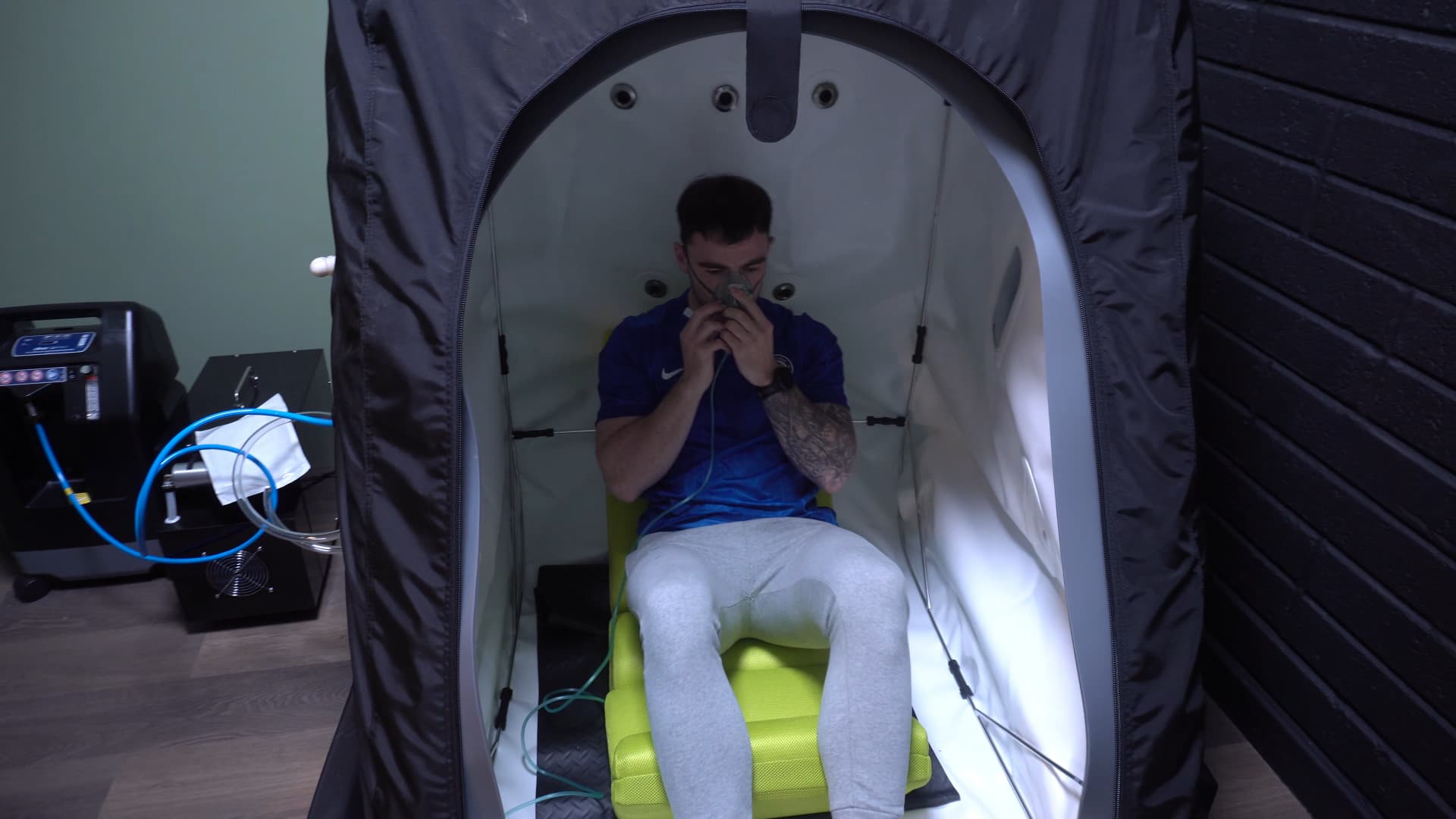 A client in the open hyperbaric chamber holding the oxygen mask to his face, the concentrator and hose alongside
