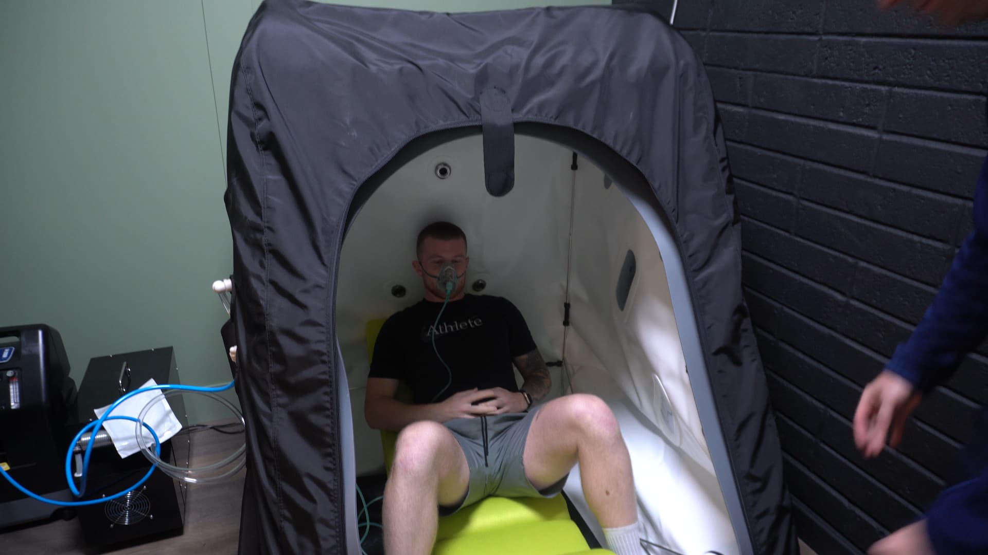 A client wearing the oxygen mask inside the chamber, a staff member's hand at the edge of the frame